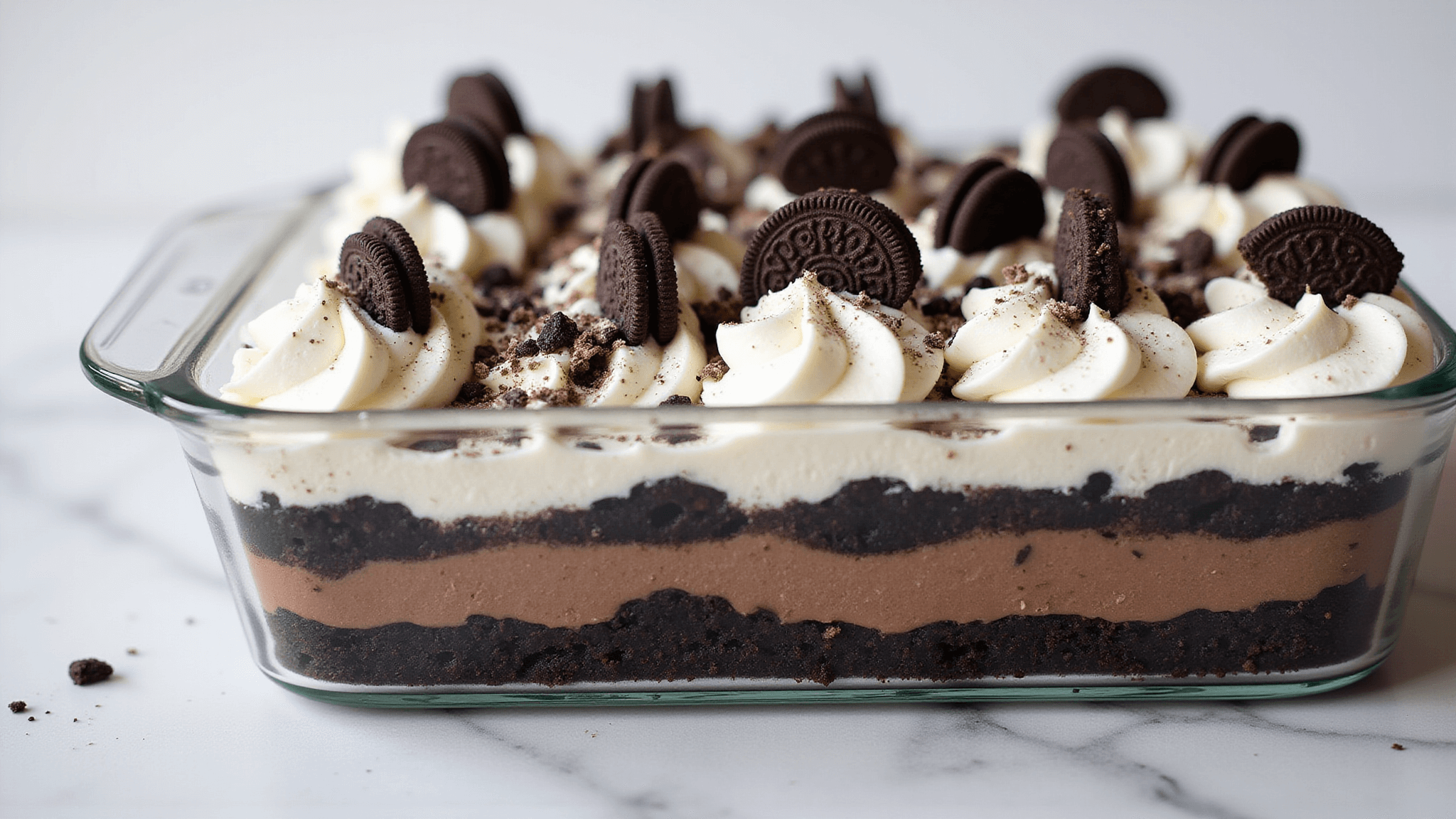 A professionally photographed no-bake Oreo dessert in a glass pan, showcasing distinct layers: Oreo crust, cream cheese, chocolate pudding, and whipped topping, garnished with crushed and whole Oreos, on a white marble background.