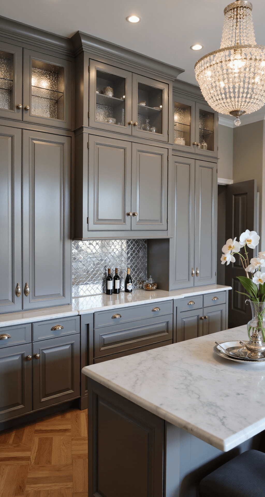 A sophisticated kitchen with grey-stained maple cabinets, a crystal chandelier casting shadows on marble countertops, polished nickel hardware, and herringbone wood floors, styled with a silver tea service, orchid arrangement, and vintage wine bottles.