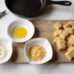 Photorealistic overhead view of a marble countertop with a professional breading station, featuring three ceramic dishes of seasoned flour, beaten eggs, and panko breadcrumbs, fresh razor clams on a cutting board, a chef's knife, and soft natural daylight casting shadows.